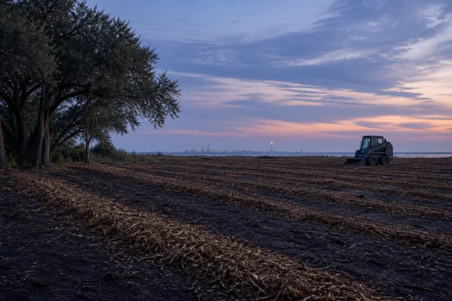 Dusk over a cleared pasture with a distant industrial skyline silhouette and a parked skid steer