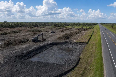 Two machines clearing a multi-acre commercial tract beside a farm-to-market highway with windrows and silt fence in place